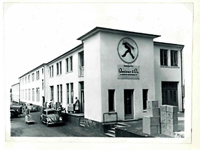 A black and white photograph of a building featuring a logo of a man walking. The structure has multiple windows and doors, with people and cars in the foreground and boxes stacked near the entrance.