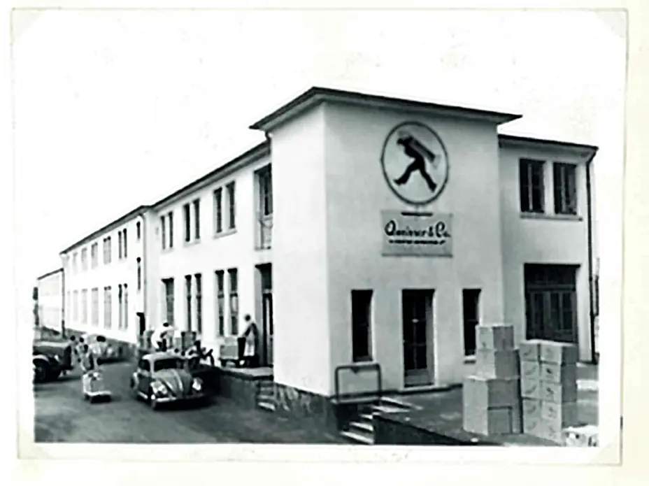 A black and white photograph of a building featuring a logo of a man walking. The structure has multiple windows and doors, with people and cars in the foreground and boxes stacked near the entrance.