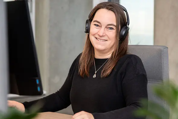 A woman with long brown hair, wearing a black sweater and headphones, is smiling while sitting at a desk with a computer monitor in front of her. Soft greenery is visible in the foreground.