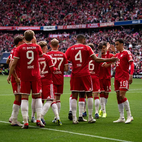 A group of FC Bayern Munich football players in red uniforms celebrates on the field, surrounded by a cheering crowd in the background. Players are engaging with each other, highlighting teamwork and camaraderie during a match.