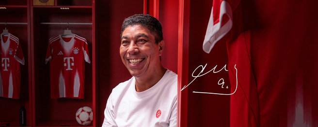 A smiling man sits in a sports locker room decorated in red, with jerseys hanging behind him. He wears a white shirt and is surrounded by soccer gear, including a ball. An autograph-like signature appears next to him.