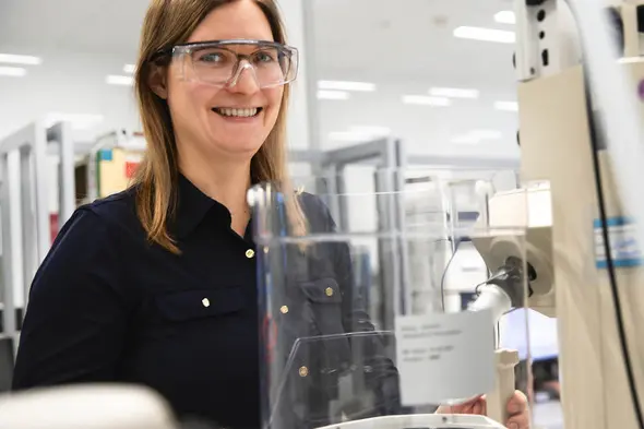 A woman with long brown hair smiles while wearing safety glasses in a laboratory setting. She stands next to a large piece of scientific equipment, appearing engaged and ready to work. The background features shelves and other lab tools.