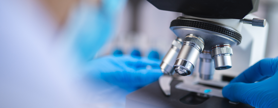 A close-up view of a microscope being handled by a person wearing blue gloves in a laboratory setting. The focus is on the microscope's lenses and the person's hands preparing a slide for observation.