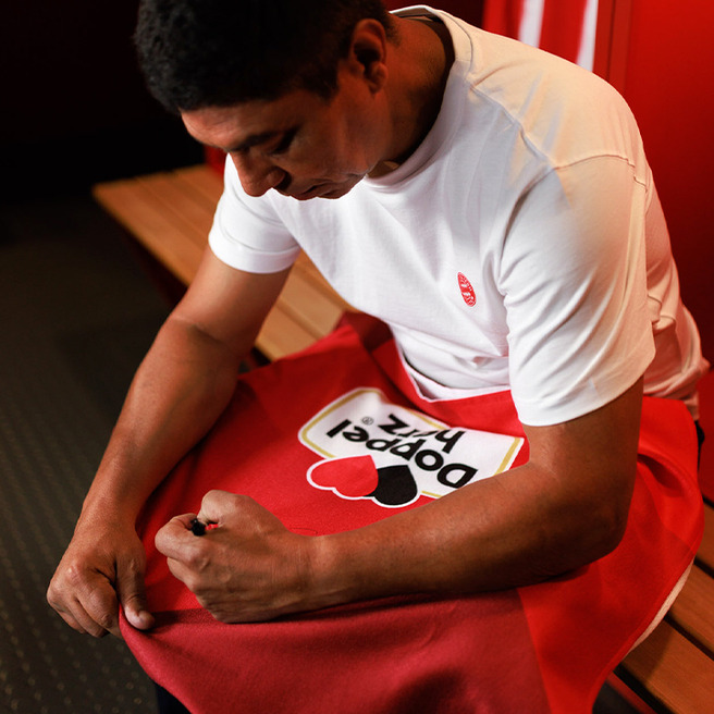 A person is seated on a bench in a locker room, looking down at a red towel with a heart design and the text "Popped Fizz." They are adjusting the towel, wearing a plain white shirt and shorts. The background is softly blurred, emphasizing the individual.