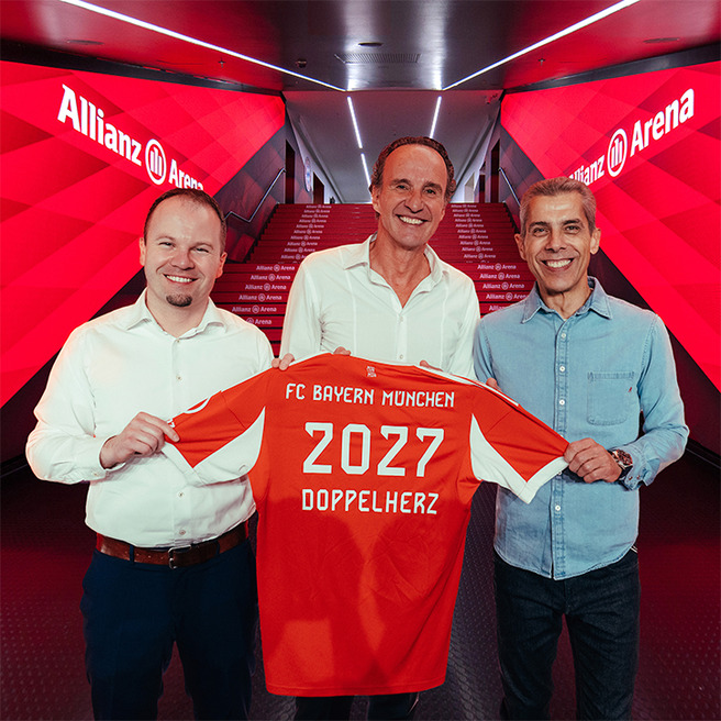 Three men stand in a brightly lit hallway at Allianz Arena, holding an orange FC Bayern München jersey. The jersey features the numbers "2027" and the word "Doppelherz." The background has vibrant red graphics, highlighting the team's branding.