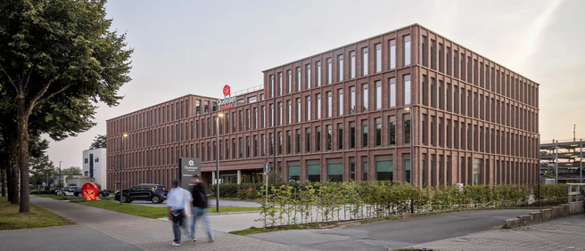 A modern brick building features large windows, surrounded by green landscaping and parking spaces. Two pedestrians walk along the sidewalk in front, and a red sculpture is visible in the foreground. The sky appears clear and the scene is set in an urban environment.