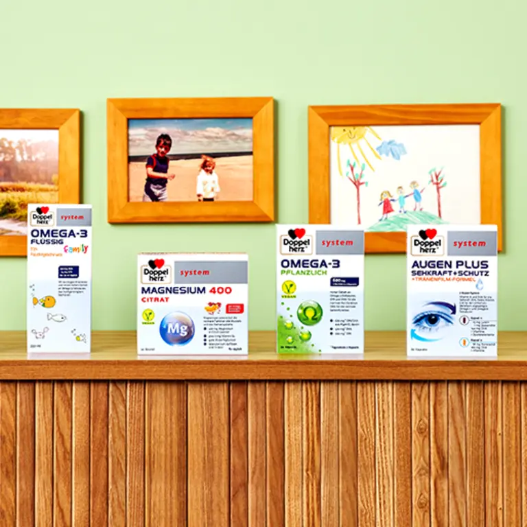A wooden shelf displays four health supplement boxes against a colorful background with framed children's drawings. The products include Omega-3 Liquid, Magnesium 400 Citrate, Omega-3 Capsules, and Augen Plus Eye Protection, all from the brand DOPPELHERZ.