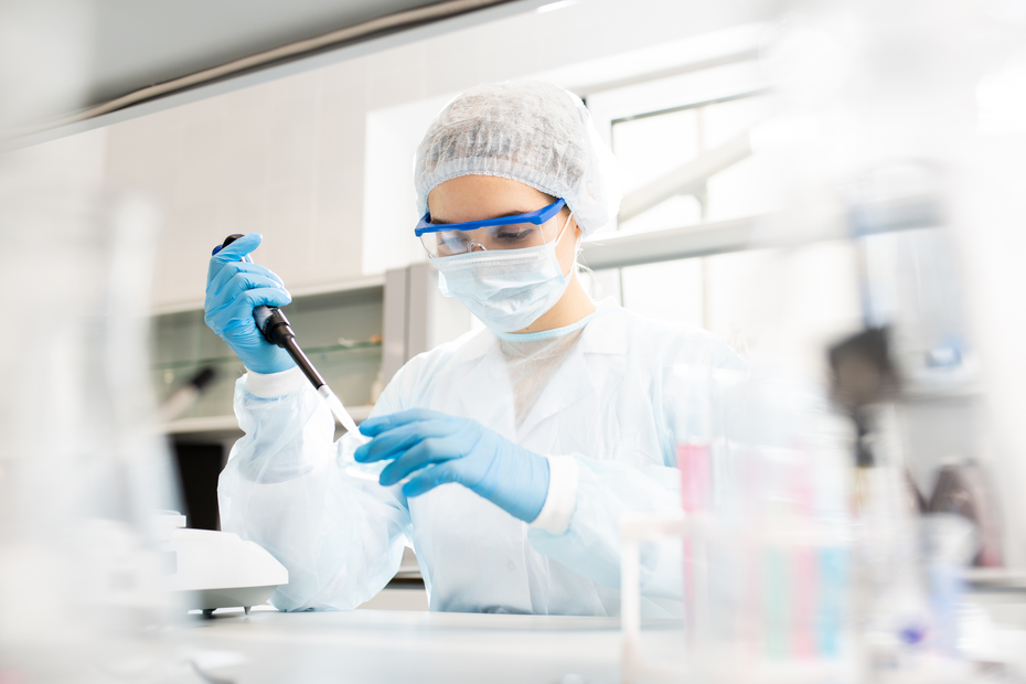 A scientist wearing a lab coat, gloves, goggles, and a mask carefully uses a pipette in a laboratory setting. The background shows laboratory equipment and glassware, indicating an environment focused on scientific research.