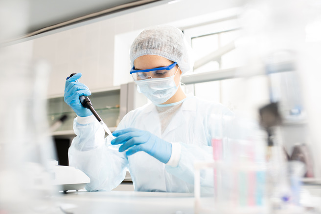 A scientist wearing a lab coat, gloves, goggles, and a mask carefully uses a pipette in a laboratory setting. The background shows laboratory equipment and glassware, indicating an environment focused on scientific research.