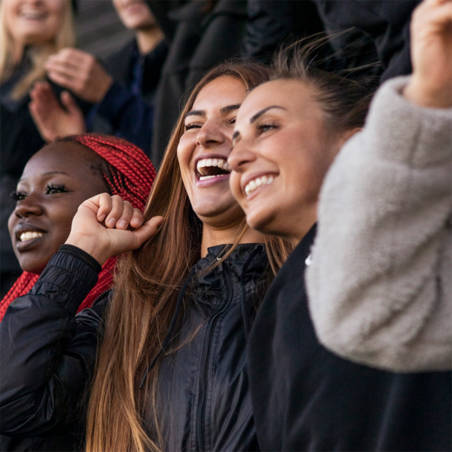 A group of three women are smiling and celebrating together, displaying joy and excitement. They are dressed casually and appear to be in a lively atmosphere, possibly during a sporting event, with their arms raised in cheer. 