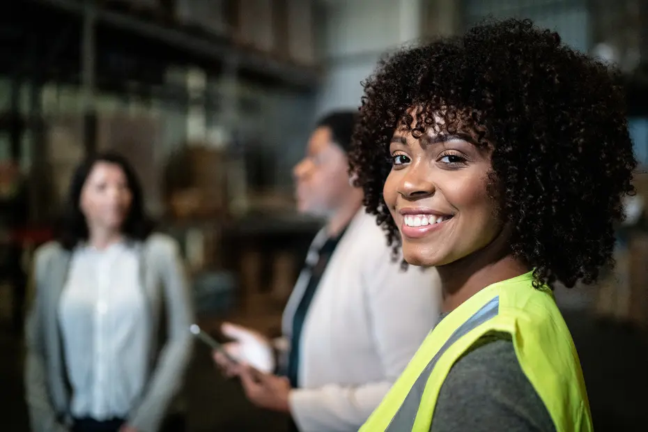 A woman with curly hair and a bright yellow safety vest smiles at the camera while two other women discuss in the background of a warehouse setting. The environment appears industrial with shelves and boxes.