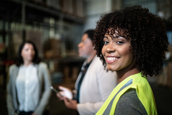 A woman with curly hair and a bright yellow safety vest smiles at the camera while two other women discuss in the background of a warehouse setting. The environment appears industrial with shelves and boxes.