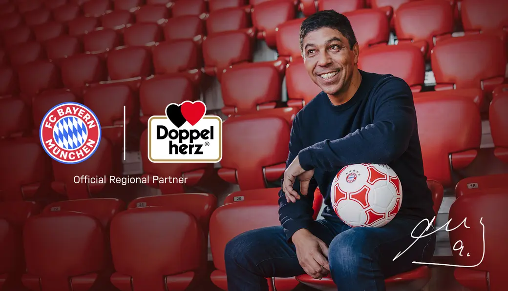 A man sits in a stadium seat, smiling and holding a soccer ball. Logos for FC Bayern München and Doppelherz appear alongside the text "Official Regional Partner" at the bottom. The background features empty seats in a stadium.