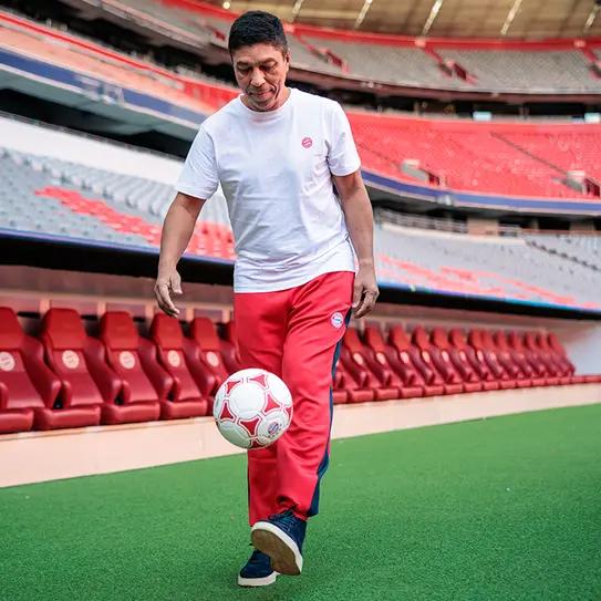 A man in a white t-shirt and red athletic pants is playing with a soccer ball on a green field inside a stadium. Empty red seats are visible in the background, highlighting the venue's space.