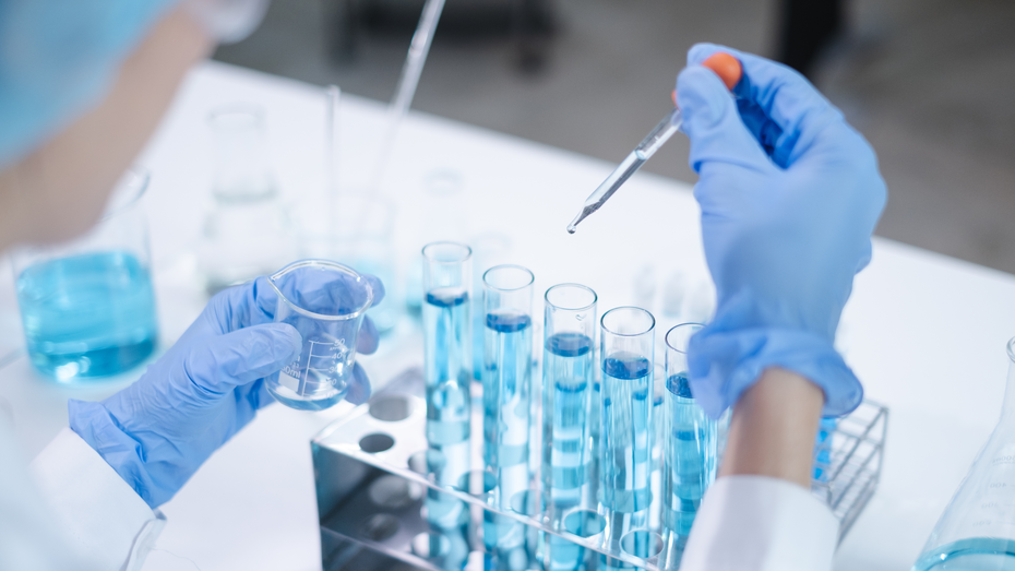 A laboratory scene showing a person's hand in a blue glove holding a pipette above test tubes filled with blue liquid. The other hand holds a small beaker, also containing blue liquid. Various glassware is visible in the background.