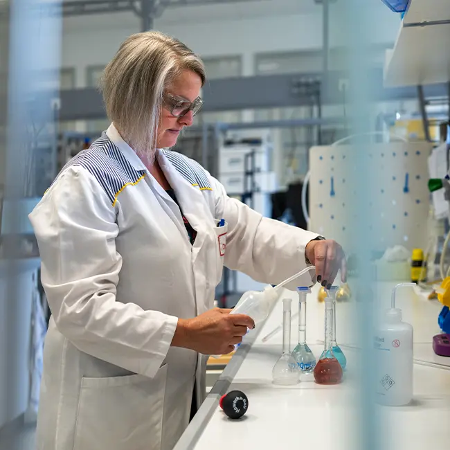 Eine Laborangestellte arbeitet mit Erlenmeyerkolben und Pipetten.  Im Hintergrund ist weitere Laborausruestung sichtbar.