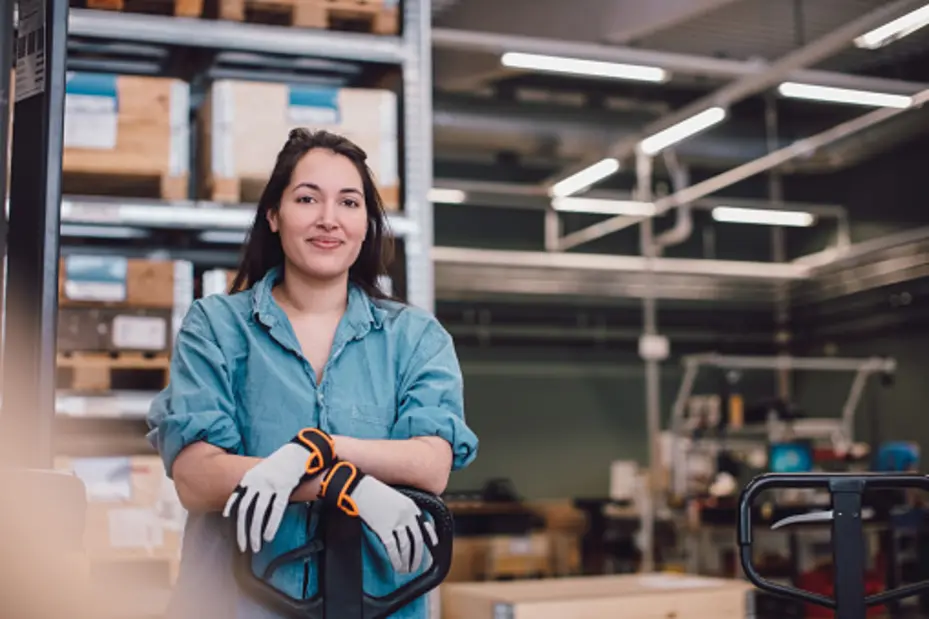 A woman wearing a light blue shirt and gloves smiles while resting her arms on a pallet jack in a warehouse setting. Behind her are shelves filled with stacked boxes and various equipment, suggesting an active work environment.