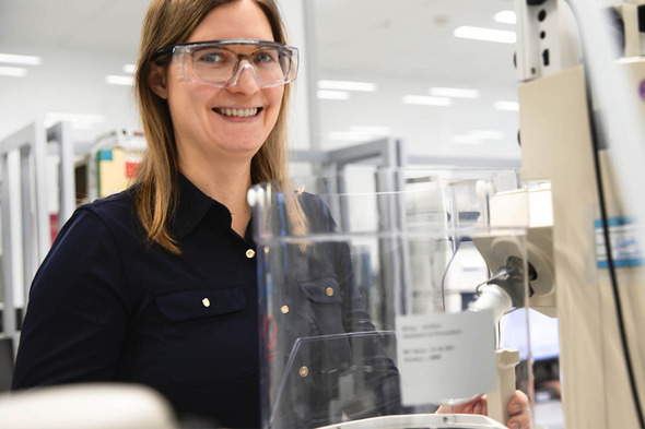 A woman with long brown hair smiles while wearing safety glasses in a laboratory setting. She stands next to a large piece of scientific equipment, appearing engaged and ready to work. The background features shelves and other lab tools.
