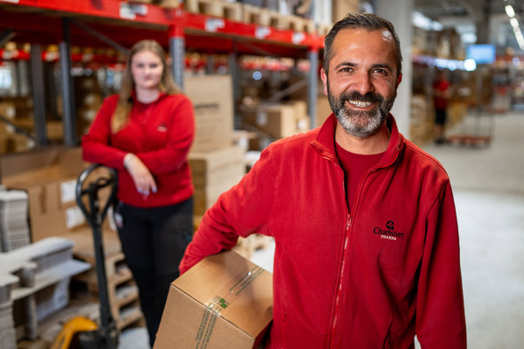 A smiling man in a red work jacket holds a box in a warehouse setting, while a woman in a similar jacket stands in the background near a pallet jack. The scene is filled with stacked boxes and shelves, indicating a busy work environment.